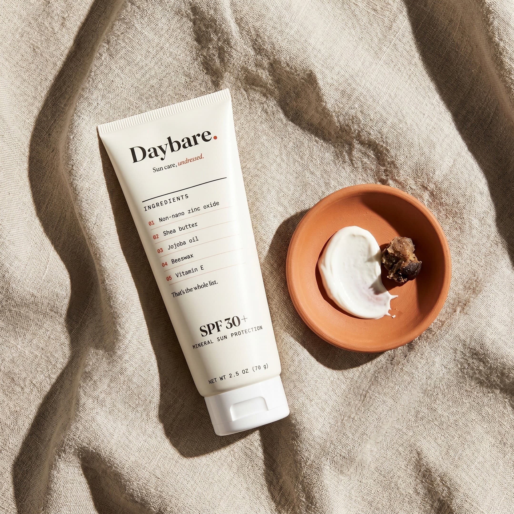 Daybare tube on raw linen beside a terracotta dish holding one smear of zinc balm